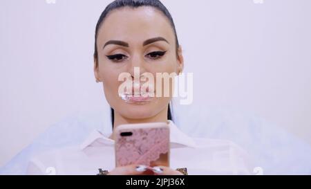 Primo piano, il paziente siede nella clinica di cosmetologia con un tampone di crema anestetico sovrapposto sulle labbra, guarda le foto sul telefono, procedura di aumento delle labbra. Foto di alta qualità Foto Stock