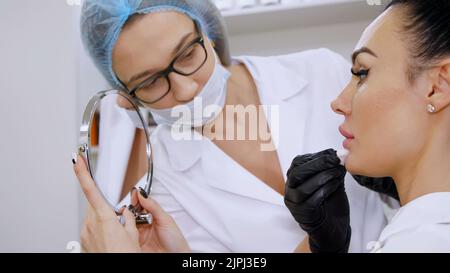 ufficio medico, bella donna che guarda nello specchio, il medico pulisce le labbra del paziente con un tovagliolo sterile, prima di iniezioni di acido ialuronico nelle labbra, pulisce la superficie con un antisettico. Foto di alta qualità Foto Stock