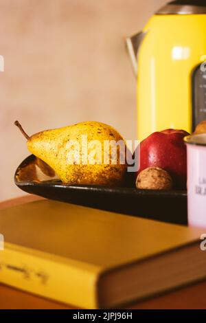 Vita morta autunnale: Tazza rosa, teiera gialla, ciotola di frutta, libro di copertina rigida sdraiato sul tavolo. Casalinghi retrò, utensili. Lettura in accogliente dolce casa atm Foto Stock