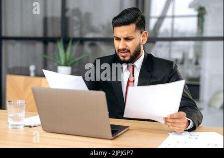 Delusione sottolineato indiano o arabo uomo d'affari, sedersi alla scrivania con computer portatile in ufficio moderno, lavora con i documenti, sconvolto con il rapporto finanziario, analizza il risultato del lavoro, si sente frustrato Foto Stock