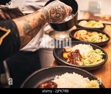 Il tocco finale. Primo piano di uno chef irriconoscibile tatuato mani che servono cibo in piatti all'interno di una cucina di un ristorante. Foto Stock