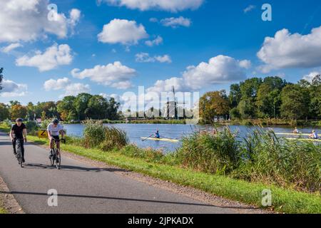 Due ciclista lungo il fiume Amstel wiyh Rieker mulino a vento in background, Amsterdam, Paesi Bassi Foto Stock