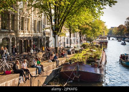I giovani si rilassano con Row Homes e House Boats lungo il canale Prinsengracht, Amsterdam, Olanda Foto Stock