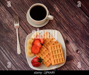 Una tazza di caffè nero e un piatto di cialde con fragole e banane su un tavolo di legno. Vista dall'alto, disposizione piatta. Foto Stock