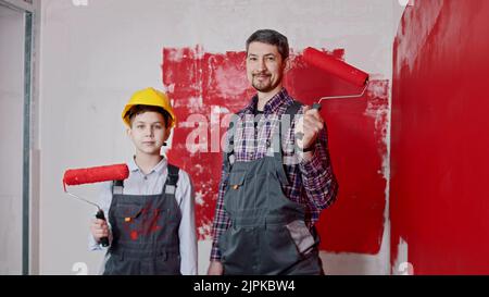 Un ragazzino e suo padre sorridente in piedi nella stanza e tenendo i rulli per la pittura a muro. Scatto medio Foto Stock