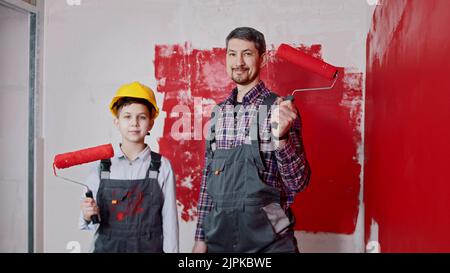 Un ragazzino e suo padre sorridente in piedi nella stanza e tenendo i rulli coperti di pittura rossa. Scatto medio Foto Stock