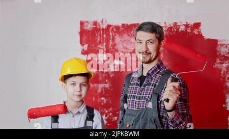 Un ragazzino e suo padre sorridente in piedi nella sala da stiro e tenendo i rulli coperti di pittura rossa. Scatto medio Foto Stock