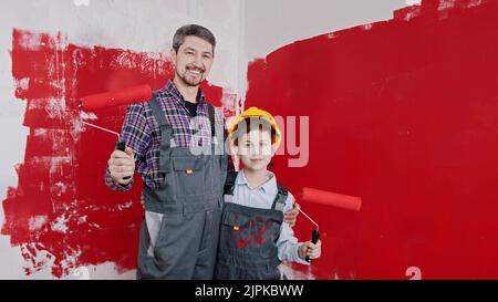 Un ragazzo e suo padre sorridente in piedi nella stanza e tenendo i rulli coperti di pittura rossa. Scatto medio Foto Stock