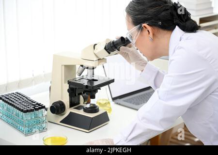 Un esperto scienziato asiatico di età femminile o un supervisore chimico medico che esamina un campione di virus sotto un microscopio mentre lavora in laboratorio. Primo piano i Foto Stock