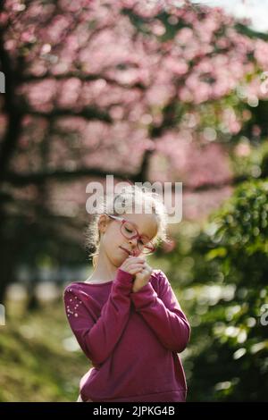 La bambina bionda ha un pelo e un odore di fiore davanti ai fiori di sakura Foto Stock