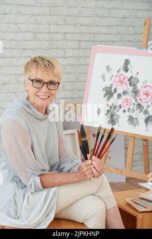 Donna anziana sorridente felice che tiene vari pennelli che usa per dipingere Foto Stock