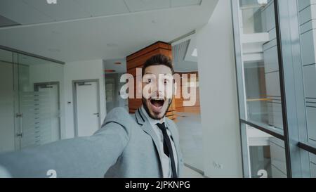 POV di un giovane uomo d'affari in tuta che ha video chat online utilizzando la fotocamera dello smartphone e parlando con i suoi colleghi in un ufficio moderno al coperto Foto Stock