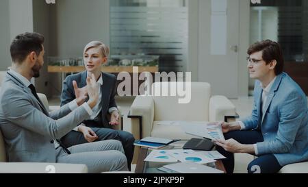 uomo d'affari che parla di relazioni finanziarie con partner commerciali femminili e maschili seduti sul divano in una moderna sala ufficio con interni Foto Stock