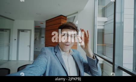 POV di giovane uomo d'affari gioioso in occhiali scattando una foto selfie e divertirsi in un ufficio moderno al coperto Foto Stock