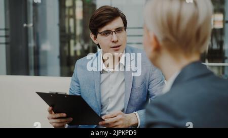 HR manager con Clifbooard intervistando giovane candidata per l'assunzione in posizione vacante seduta sul divano in moderno ufficio interno Foto Stock
