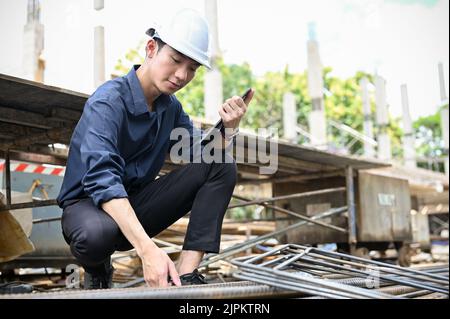 Bello e professionale giovane ingegnere civile asiatico maschio in hardHat bianco che controlla la barra di acciaio nel cantiere. Foto Stock