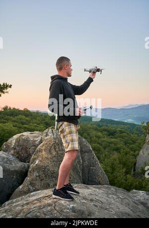 Uomo che utilizza il drone tramite telecomando. Uomo che usa il drone al tramonto per foto e video making mentre si trova in cima a un masso alto in montagna. Foto Stock