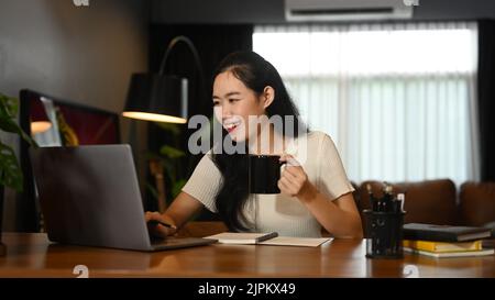 Bella donna asiatica che tiene una tazza di caffè e utilizza un computer portatile su una scrivania di legno Foto Stock