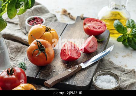 Pomodori crudi e coltello su tagliere di legno circondato da ingredienti sale foglie di basilico e olio d'oliva intorno. Sfondo rustico cucina. Orisontale Foto Stock