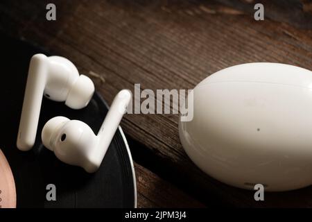 Le cuffie wireless e la custodia di ricarica si trovano su un tavolo di legno su un disco in vinile, musica e hobby Foto Stock