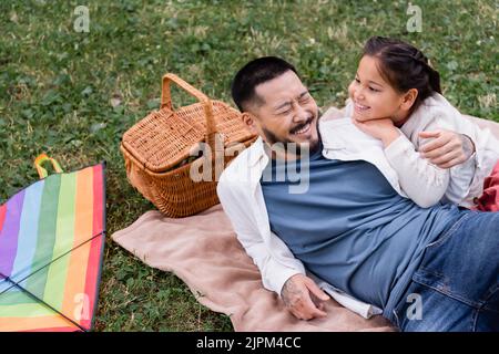 Felice papà asiatico che si trova vicino a figlia e cesto di wicket nel parco Foto Stock