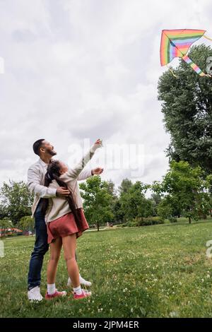 Vista laterale di sorridente padre asiatico che abbraccia figlia con aquilone volante nel parco Foto Stock