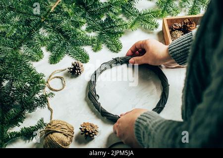 Donna in maglia grigia maglione fare una corona di abete di natale. Decorazione di Natale per gli interni domestici. Passo dopo passo, processo. Fai da te, artigianato, hobby conce Foto Stock