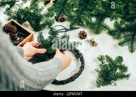 Donna in maglia grigia maglione che fa la corona di abete di natale. Decorazione di Natale per gli interni domestici. Passo dopo passo, processo. Fai da te, artigianato, concetto di hobby Foto Stock