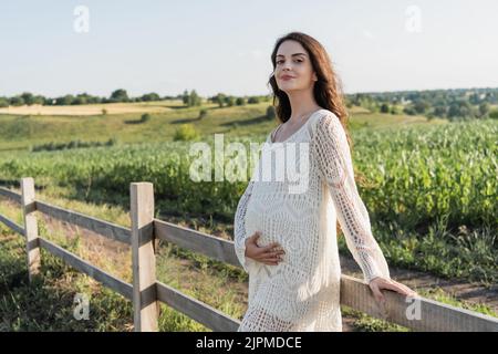 donna incinta felice in vestito bianco openwork in piedi vicino recinto di legno in campo Foto Stock