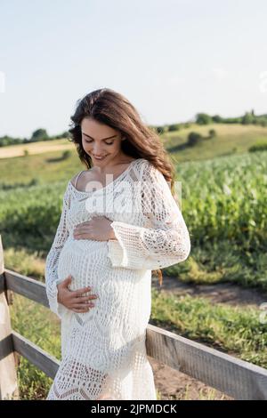 bella donna incinta in vestito bianco che tocca il ventre e sorridente all'aperto Foto Stock