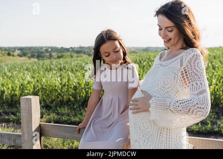 ragazza in abito rosa seduta su recinzione di legno vicino felice e madre incinta Foto Stock