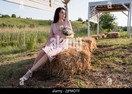 intera lunghezza di donna in abito rosa seduta su pagliaio con bouquet di fiori Foto Stock