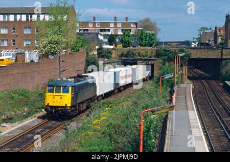 Una locomotiva elettrica di classe 85 numero 85114 che lavora un freightliner ben caricato passa dietro le piattaforme della stazione di Caledonian Road & Barnsbury sulla North London Line. 14th maggio 1991. Foto Stock