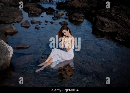 Vista laterale di una giovane femmina graziosa e barefooted con lunghi capelli scuri in vestito bianco seduto sulla pietra in mare e abbracciando le stelle marine nella natura Foto Stock