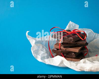 Pila di cioccolato avvolta con un nastro rosso su sfondo blu, spazio copia. Concetto di regalo dolce, regalo piacevole Foto Stock