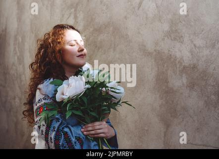 Ragazza con gli occhi chiusi tiene bouquet di peonie. Donna in camicia ricamata e con lunghi capelli ricci rossi contro pareti in cemento. Tasto scuro foto. Giornata dell'indipendenza dell'Ucraina. Spazio di copia. Messa a fuoco morbida Foto Stock