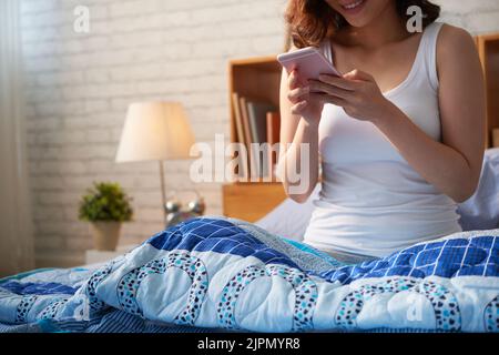 Immagine ritagliata di una giovane donna seduta a letto e che controlla il telefono al mattino Foto Stock