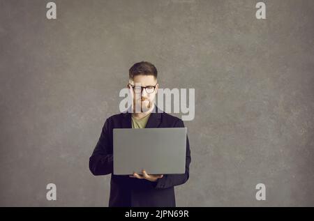 Programmatore IT maschile serio e concentrato in piedi con un portatile in mano su uno sfondo grigio. Foto Stock