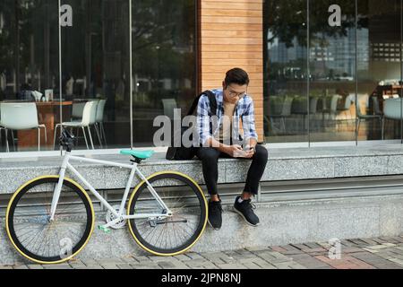 Ritratto completo di un bel ciclista asiatico seduto di fronte alla moderna caffetteria e navigando su Internet sullo smartphone Foto Stock