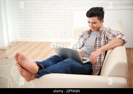 Un bel giovane vietnamita sorridente che riposa sul divano con un computer portatile Foto Stock