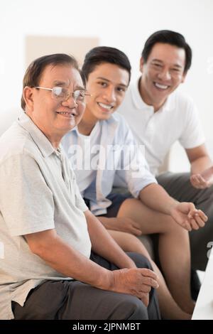 Allegro nonno vietnamita, padre e figlio seduti sul divano e guardando la macchina fotografica Foto Stock