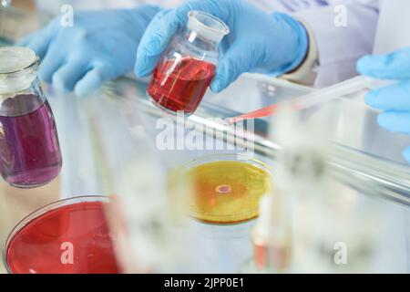 Primo piano di uno scienziato irriconoscibile che indossa guanti di gomma utilizzando una pipetta mentre si miscelano due liquidi in una capsula di Petri, vetreria da laboratorio in primo piano Foto Stock