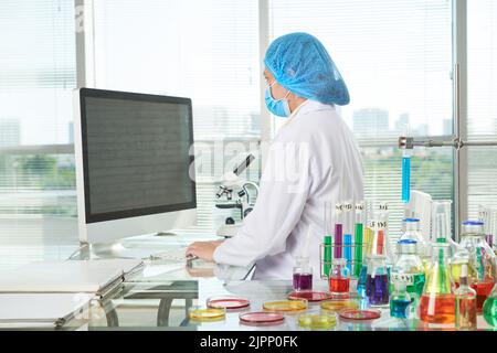 Vista del profilo di una ricercatrice femminile seduta di fronte al moderno computer e prendendo appunti necessari dopo il successo del promettente esperimento Foto Stock