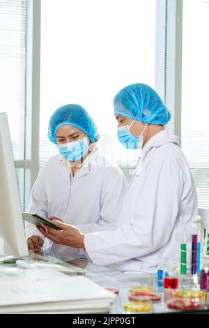 Gruppo di ricercatori che lavorano sodo indossando maschere mediche e camici bianchi seduti al banco di laboratorio e studiando i risultati di esperimenti condotti con l'aiuto di tablet digitale Foto Stock