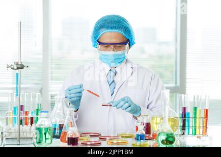 Operatore medico altamente professionale che indossa abiti bianchi e occhiali di protezione avvolti in ufficio mentre si trova al banco da laboratorio, con foto in verticale Foto Stock