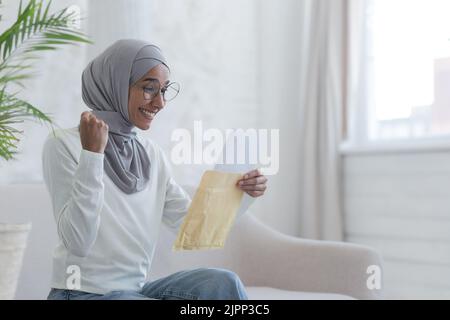 Una giovane bella donna musulmana legge una felice lettera casa studente arabo ha ricevuto conferma dall'università superato con successo l'esame una donna in un hijab è seduta su un divano Foto Stock