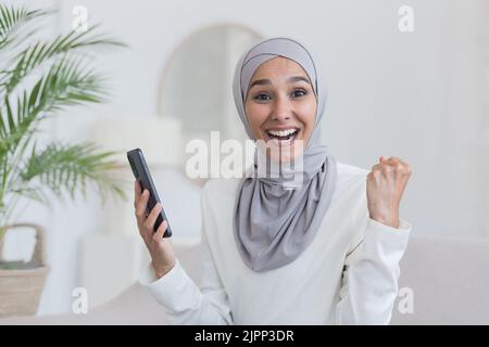 Ritratto di giovane bella donna in hijab, arabo musulmano donna guardando la macchina fotografica e sorridente seduto sul divano in soggiorno, tenendo il telefono in mano felice successo vittoria e buone notizie Foto Stock