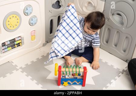 Bambino ragazzo di 11 mesi a casa di sollevamento panno per trovare giocattolo nascosto Piaget oggetto Permanence serie #2 Foto Stock
