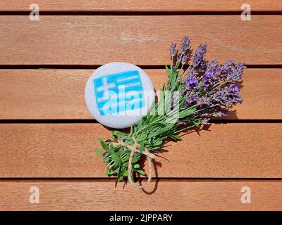 Bandiera di paese della Grecia dipinta sulla pietra di mare con mazzo di fiori freschi di lavanda su fondo di legno tavola. Foto Stock