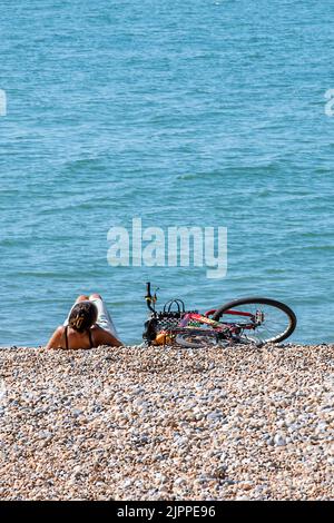 lady sdraiata su una spiaggia di ghiaia rocciosa sulla ghiaia accanto alla sua bicicletta nel caldo sole estivo prendendo il sole durante un'onda di caldo estivo, donna in spiaggia. Foto Stock